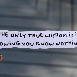 May include: A white sticker with the quote "THE ONLY TRUE WISDOM IS IN KNOWING YOU KNOW NOTHING." in black text. The sticker is held by a hand with a black and white patterned manicure.