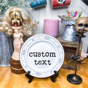 May include: A white ceramic plate with a floral design and the text "custom text" in black. The plate is on a black stand and is surrounded by various decorative items, including a doll with a mask, a candle, and a metal figurine.