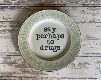 Może przedstawiać: Biały ceramiczny talerz ze złotym obramowaniem i zieloną kwiatową obwódką. Na talerzu widnieje napis "say perhaps to drugs" czarnymi literami. Talerz umieszczony jest na zniszczonej, pomalowanej na biało drewnianej powierzchni.
