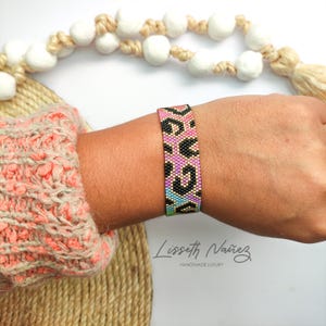Puede incluir: Un brazalete de cuentas con un diseño de estampado de leopardo en rosa, negro, morado, azul y verde. El brazalete se lleva en la muñeca. El fondo incluye un suéter beige y elementos decorativos.