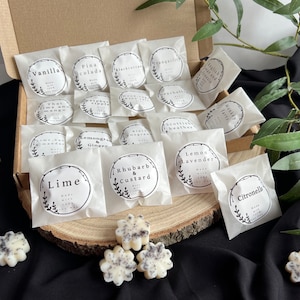 May include: A collection of small, white wax melts in individual paper bags. Each bag has a black and white label with a different scent name, such as "Vanilla", "Lime", and "Rhubarb & Custard".