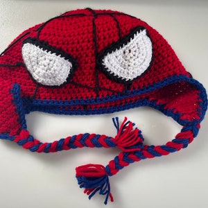 May include: A handmade crochet hat resembling Spider-Man. The hat is predominantly red with black webbing and white eye shapes. Blue trim and braided ear flaps with red and blue tassels complete the design.