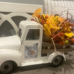 May include: White toy truck with a wooden bed filled with fall-themed decorations, including faux pumpkins, corn, and leaves. The truck has a sign that reads "Pumpkin Patch Farm Fresh Pick Your Own".