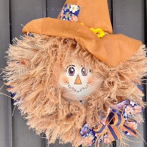 May include: A brown scarecrow wreath with a straw-like texture, a painted face, and a brown hat with blue and white floral fabric accents. A blue and orange ribbon with floral print is tied around the wreath.