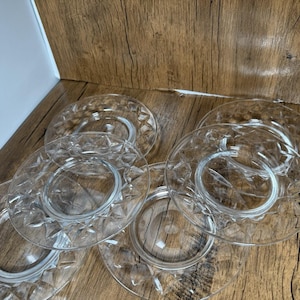May include: A collection of clear glass plates with a diamond pattern. The plates are arranged on a wooden surface, showcasing their transparent quality. The plates have a circular design with a raised center.