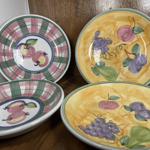 May include: Four ceramic plates with fruit designs. Two plates have a plaid pattern with apple illustrations. The other two plates have a yellow background with grapes, pears, and apple illustrations. The plates have a white rim.