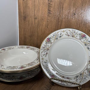 May include: A set of white porcelain dinnerware with a floral and gold rim design. The set includes plates and bowls, perfect for serving meals. The plates and bowls are stacked on a wooden surface.