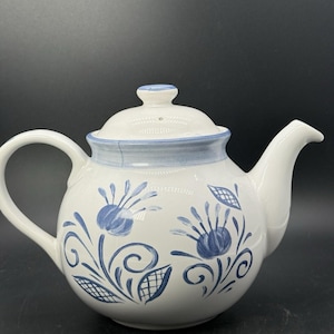 May include: A white ceramic teapot featuring a hand-painted blue floral pattern with swirling stems and leaves. The teapot includes a matching lid with a small knob, a curved handle, and a tapered spout, all decorated with a light blue rim accent.