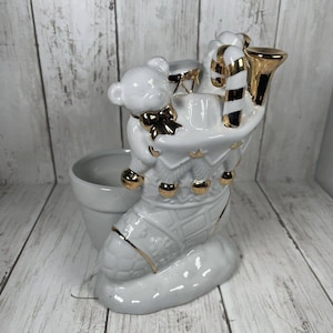 May include: White ceramic Christmas stocking planter with a gold trim and a teddy bear holding a drum. The stocking has a candy cane and a trumpet sticking out of the top.