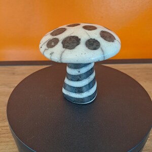 May include: A white ceramic mushroom with black polka dots and black and white stripes on the stem. The mushroom is sitting on a black circular base.