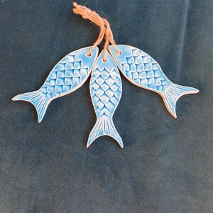 May include: Three blue ceramic fish ornaments, linked by twine. Each fish features a scale pattern and a curved tail. The ornaments are set against a dark blue backdrop. The fish are approximately 15 cm long.