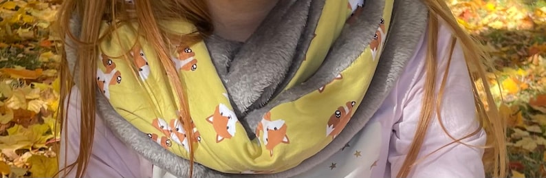 May include: A yellow infinity scarf with a gray fleece lining. The scarf features a repeating pattern of cartoon foxes.