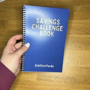 May include: A blue spiral-bound notebook titled "SAVINGS CHALLENGE BOOK" with silver lettering. The notebook is held by a hand with white nail polish. The text "DebtFreePanda" is printed at the bottom of the cover. The notebook is on a wooden surface.