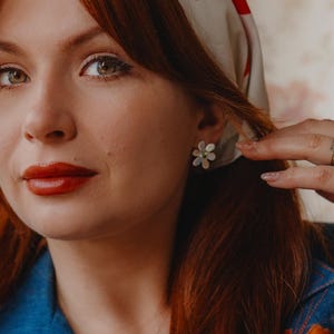 May include: A woman with red hair and red lipstick wears a white scarf with red floral designs. She is wearing a denim jacket with orange floral embroidery and flower-shaped earrings. Gold rings adorn her fingers.