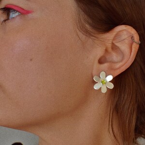 May include: Silver daisy stud earrings with a green center stone. The earrings are worn on the ear of a person with brown hair and pink eyeliner.