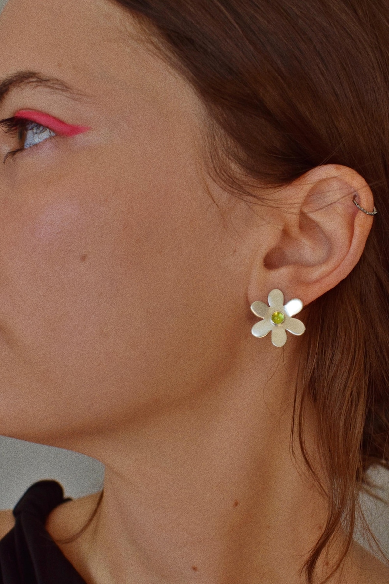 May include: Silver daisy stud earrings with a green center stone. The earrings are worn on the ear of a person with brown hair and pink eyeliner.