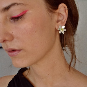 May include: Silver daisy earrings with a small yellow center. The earrings are worn on the ear of a person wearing a black top. Bright pink eyeliner is visible on the upper eyelid.