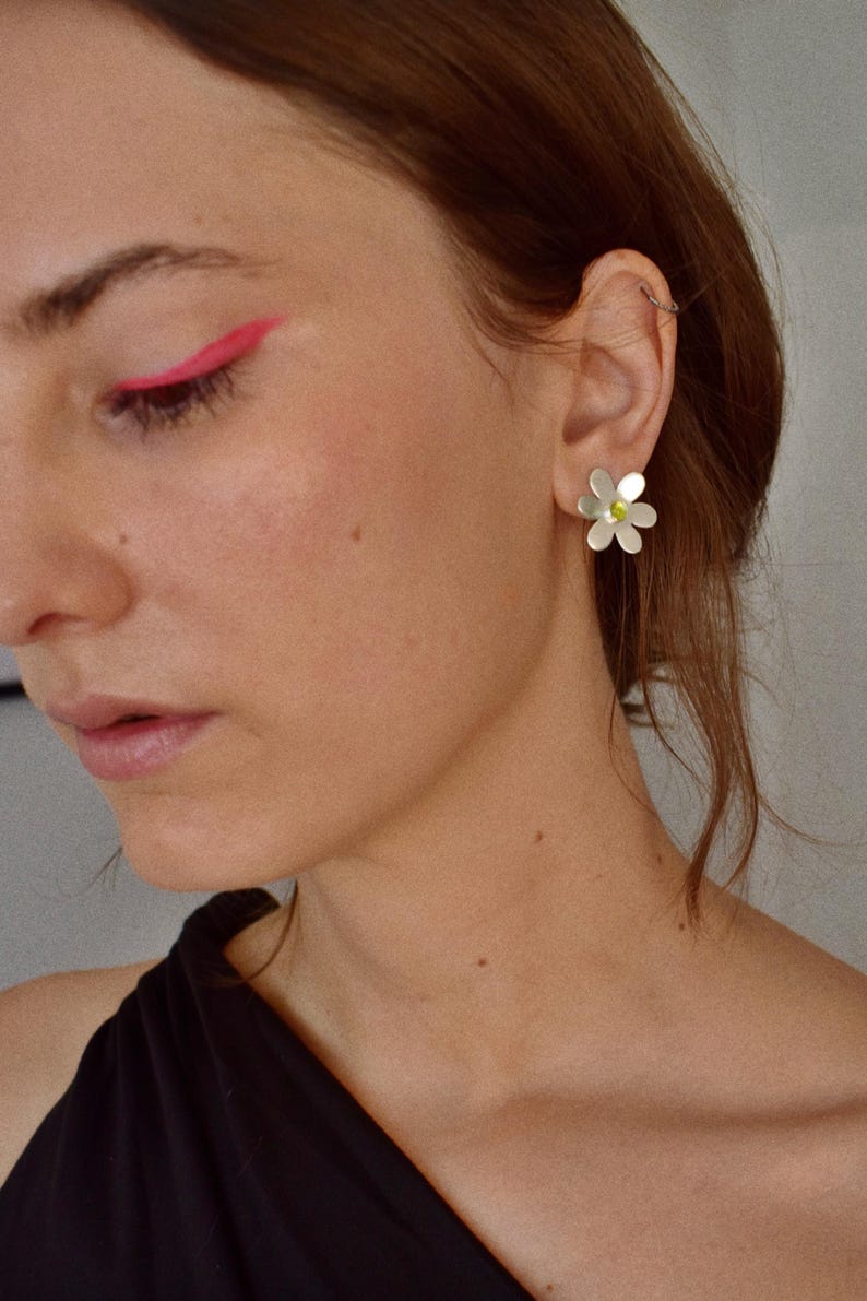 May include: Silver daisy earrings with a small yellow center. The earrings are worn on the ear of a person wearing a black top. Bright pink eyeliner is visible on the upper eyelid.