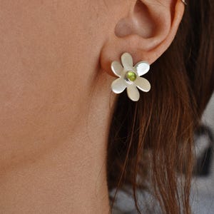 May include: Silver daisy stud earrings with a green gemstone center. The earrings are worn on the ear of a person with brown hair. The earrings have a simple, elegant design.
