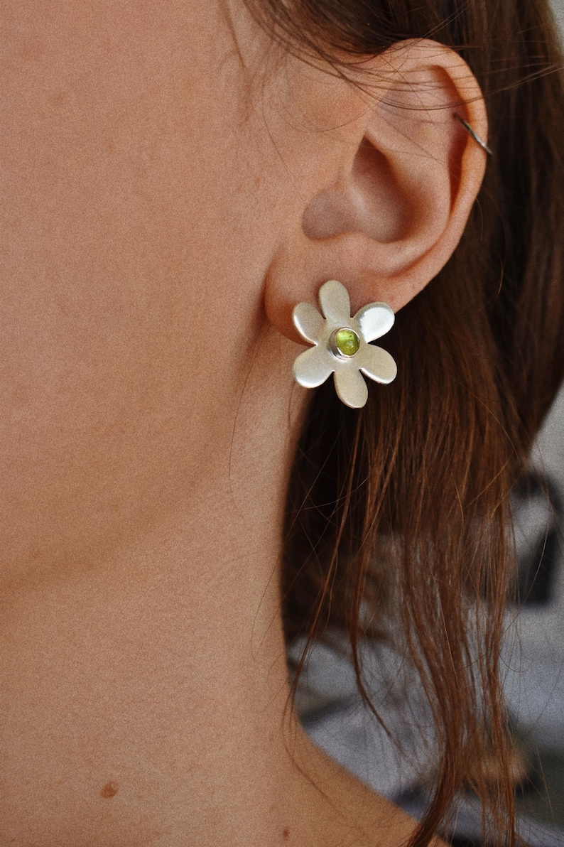May include: Silver daisy stud earrings with a green gemstone center. The earrings are worn on the ear of a person with brown hair. The earrings have a simple, elegant design.