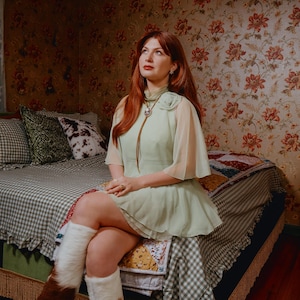 May include: A person wearing a light green dress with sheer sleeves and white fur-lined boots sits on a bed. The bed has a checkered bedspread and decorative pillows. The room has floral wallpaper and a vintage aesthetic.