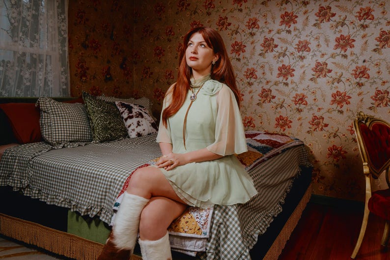May include: A person wearing a light green dress with sheer sleeves and white fur-lined boots sits on a bed. The bed has a checkered bedspread and decorative pillows. The room has floral wallpaper and a vintage aesthetic.