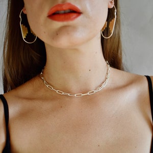 May include: A silver chain necklace with a simple, linked design. The necklace is worn by a person with long brown hair and red lipstick.