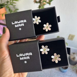 May include: Two pairs of flower-shaped earrings with a silver finish and a green center stone, displayed in black boxes. The boxes are labeled "LAURA MAR" with a star symbol.