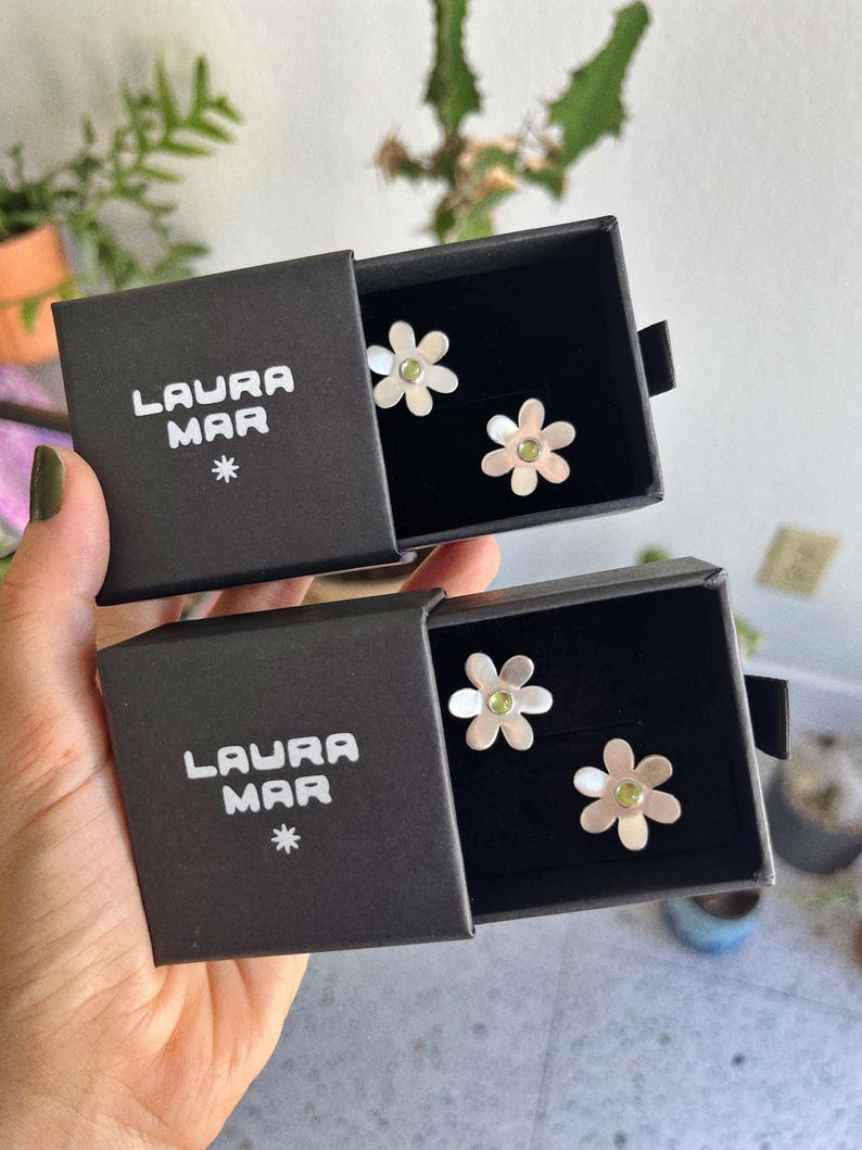 May include: Two pairs of flower-shaped earrings with a silver finish and a green center stone, displayed in black boxes. The boxes are labeled "LAURA MAR" with a star symbol.