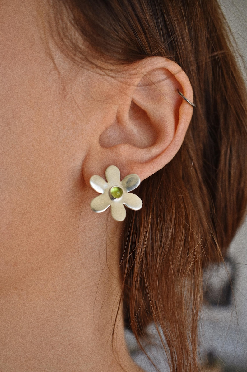 May include: Silver flower stud earrings with a small, round, green gemstone center. The earrings are worn on the ear of a person with brown hair. The earrings are a floral design.
