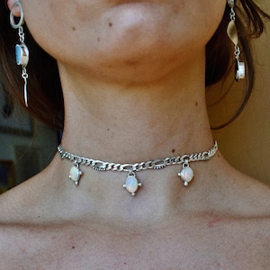 Pode incluir: Um colar gargantilha de corrente prateada com três pingentes ovais de opala. O colar está sendo usado por uma pessoa.