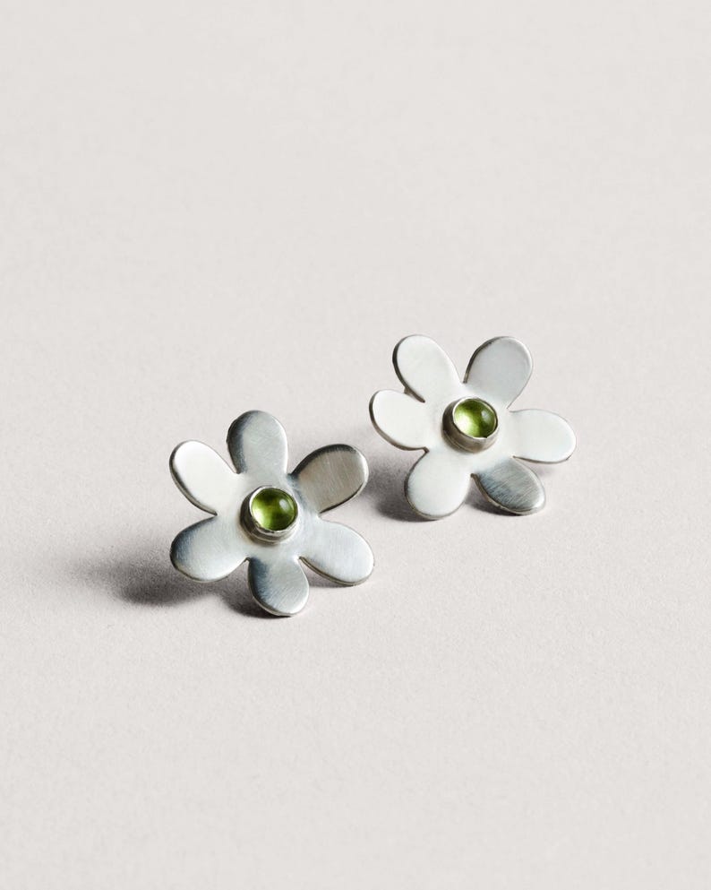 May include: A pair of silver-toned flower stud earrings. Each earring features a daisy-like design with five petals and a small, round, green gemstone in the center. The earrings are displayed on a light background.