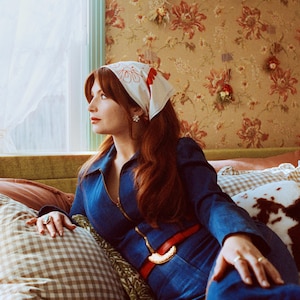 May include: A woman wearing a blue denim jumpsuit, red belt, and a white bandana with red floral designs. She is sitting on a bed with patterned pillows, with floral wallpaper in the background.