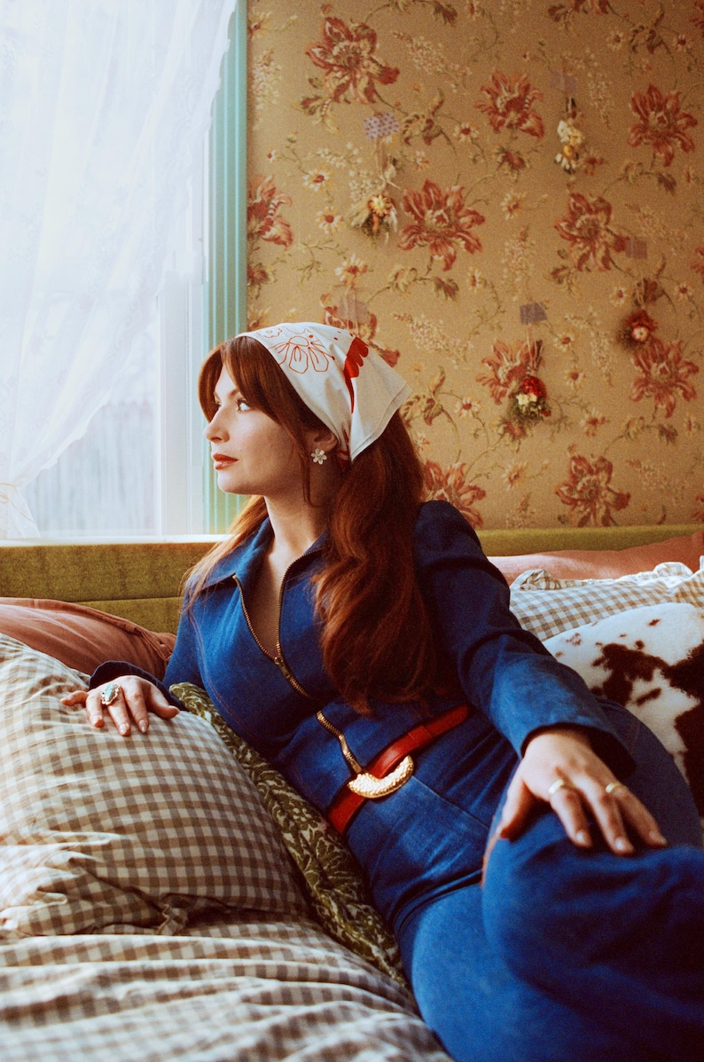 May include: A woman wearing a blue denim jumpsuit, red belt, and a white bandana with red floral designs. She is sitting on a bed with patterned pillows, with floral wallpaper in the background.