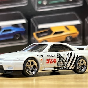 May include: A white die-cast model car with chrome wheels and a rear spoiler. The car features a Godzilla graphic with Japanese text and claw marks on the side. The Hot Wheels logo is visible.