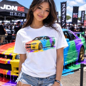 May include: A white short-sleeve t-shirt featuring a graphic print of a sports car painted in a rainbow color scheme with a dripping paint effect. The car design includes yellow, blue, green, and purple sections with bronze-colored wheels.