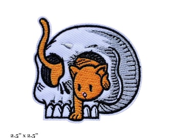 May include: An embroidered iron-on patch featuring a white skull with a small orange cat peeking out from inside. The patch is 2.5 inches by 2.5 inches, or 6 centimeters by 6 centimeters.