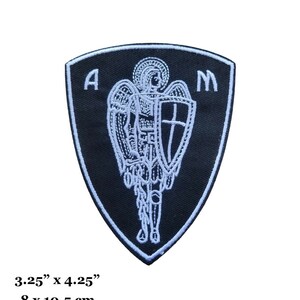 Pode incluir: Etiqueta bordada preta com um anjo branco segurando uma espada e um escudo. O anjo tem asas e uma auréola. A etiqueta tem a forma de um escudo e tem as letras "A" e "M" de cada lado. A etiqueta mede 8 cm por 10,5 cm.