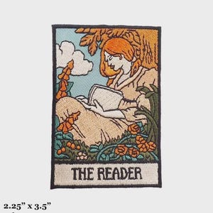 May include: Embroidered iron-on patch featuring a woman reading a book in a garden setting. The patch is surrounded by a black border and has the text "THE READER" at the bottom.