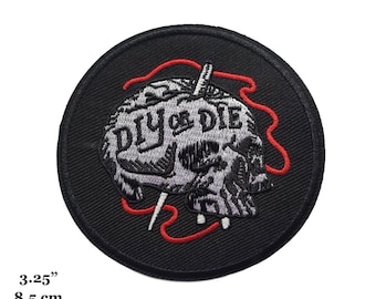 Puede incluir: Parche bordado en blanco y negro con una calavera y el texto "DIY or DIE" en un círculo. La calavera está cosida con hilo rojo.