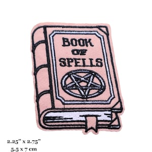 May include: A pink embroidered iron-on patch featuring a closed book with the text "Book of Spells" and a pentagram symbol. The patch measures 2.25 inches by 2.75 inches.