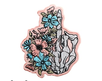 May include: An embroidered iron-on patch featuring a skeleton hand holding a bouquet of pink and blue flowers. The patch is 2.75 inches by 3.25 inches and 6.5 cm by 8.5 cm.
