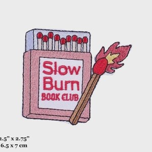 Op de afbeelding: Roze en rode geborduurde strijk-op patch van een luciferdoosje met de tekst "Slow Burn Book Club" en een brandende lucifer. De patch is 6,5 cm bij 7 cm.