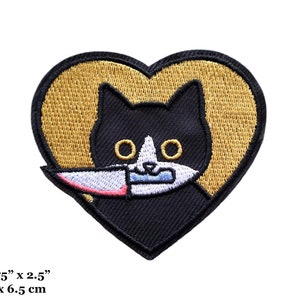 May include: Embroidered patch featuring a black cat with a white muzzle and eyes, holding a knife in its mouth. The cat is set against a gold heart-shaped background. The patch measures 2.75" x 2.5".