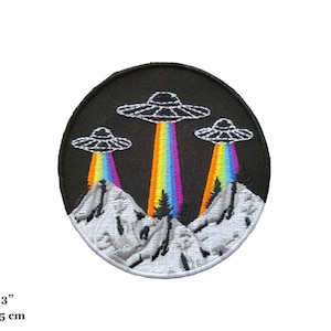 May include: A round embroidered patch with three flying saucers over a mountain range. The saucers are white with black outlines and are emitting rainbow beams of light. The mountains are white with gray outlines. The patch is 3 inches in diameter.