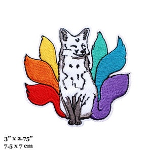 Op de afbeelding: Geborduurde strijkpatch met een witte vos met regenboogkleurige staarten. De vos zit met zijn poten bij elkaar en heeft een vrolijke uitdrukking. De patch is 7,5 cm bij 7 cm.