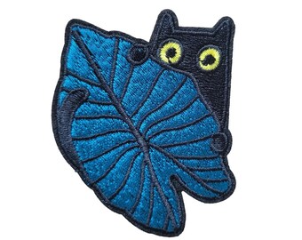 Può includere: Gatto nero che spunta da dietro una grande foglia tropicale blu. Il gatto ha gli occhi gialli. La toppa misura 2,5 pollici per 3,25 pollici, o 6 per 8 centimetri.