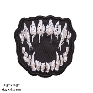 Könnte beinhalten: Ein schwarzer, gestickter Aufnäher mit einem stilisierten offenen Mund mit scharfen, weißen Zähnen. Der Aufnäher misst 6,5 cm x 6,5 cm und hat einen gewellten Rand. Die Zähne haben ein strukturiertes Aussehen, vor schwarzem Hintergrund.