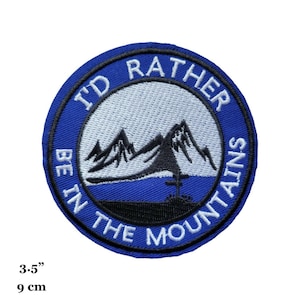 Puede incluir: Un parche bordado azul y blanco con el texto "I'd rather be in the mountains" y una representación de montañas. El parche tiene un diámetro de 9 cm.