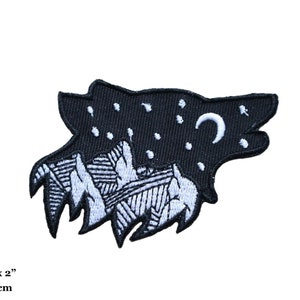 May include: Black and white embroidered patch of a wolf's head with a crescent moon and stars. The wolf's head is filled with a mountain range design. The patch measures 3.25 inches by 2 inches.